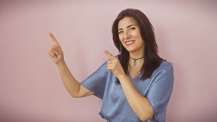 Fototapeta premium Smiling mature hispanic woman pointing upwards with both hands, against a soft pink background.