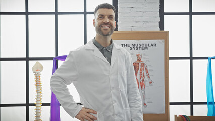 Handsome hispanic man in labcoat smiling in a physiotherapy clinic with anatomy poster.