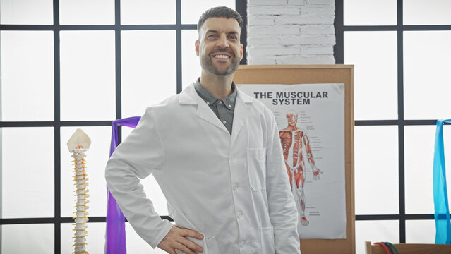 Handsome hispanic man in labcoat smiling in a physiotherapy clinic with anatomy poster.