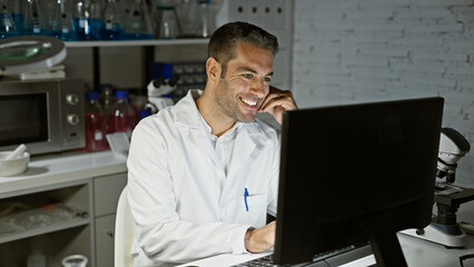 Hispanic man in lab coat smiling while working on computer in a science laboratory setting