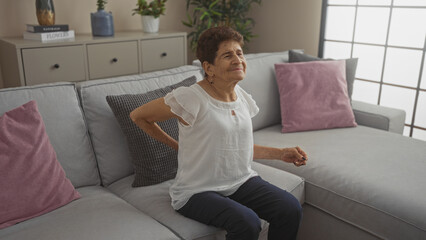 An elderly hispanic woman with short hair sits on a couch in a living room, experiencing back pain in a home environment.