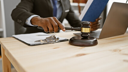 African american man in suit using gavel at desk with laptop in office setting conveying law, authority, and professionalism.