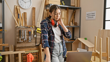 Hispanic woman with headphones stands in a carpentry workshop surrounded by tools and wood.