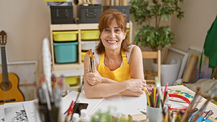 Smiling mature woman artist in yellow apron indoors studio with brushes