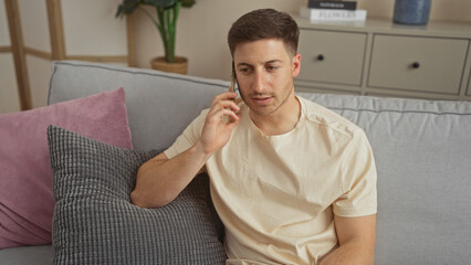 Handsome young hispanic man in a living room talking on the phone while sitting on a couch indoors at home.