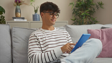 Fototapeta premium Handsome young hispanic man relaxing indoors using a tablet in a modern living room, surrounded by cozy decor and houseplants.