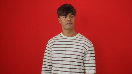 Portrait of a handsome young hispanic man wearing a striped shirt against a vibrant red background wall, isolated and looking to the side.