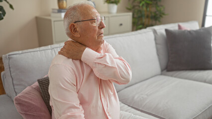 Senior, grey-haired man sitting indoors on a couch at home holding his neck in pain, suggesting discomfort or a common health issue related to aging in an interior living room setting.