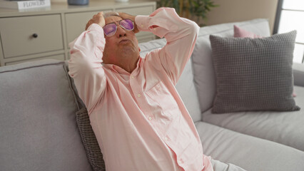 Middle-aged caucasian man feeling stressed in a cozy living room with grey-haired and wearing sunglasses indoors seated on a couch.