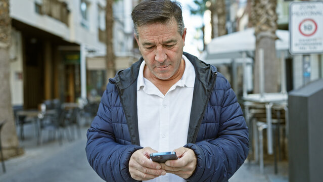 Middle-aged man in urban setting uses smartphone on city street outside a coffee shop.