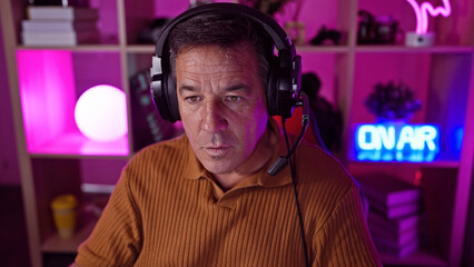 A focused middle-aged man wearing headphones in a colorful lit gaming room at night.