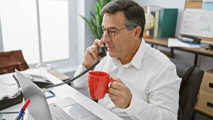 A professional middle-aged man is engaged in a conversation on the phone in a modern office setting, holding a red mug. - Powered by Adobe