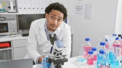 A young adult man with curly hair works in a laboratory, studying samples through a microscope, surrounded by flasks and beakers.