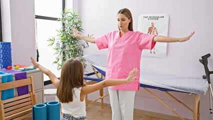Pediatrician in pink demonstrating exercise to young girl patient in white in a therapy room with...