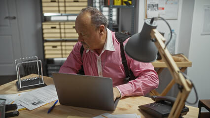 Mature bald man in a detective office analyzing evidence with laptop and paperwork.
