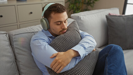 Handsome young hispanic man with a beard relaxing indoors on a comfortable couch, wearing headphones and hugging a pillow in a modern living room.