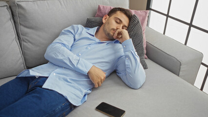 A handsome young hispanic man with a beard is sleeping on a sofa in the living room of his apartment, with his smartphone placed beside him.