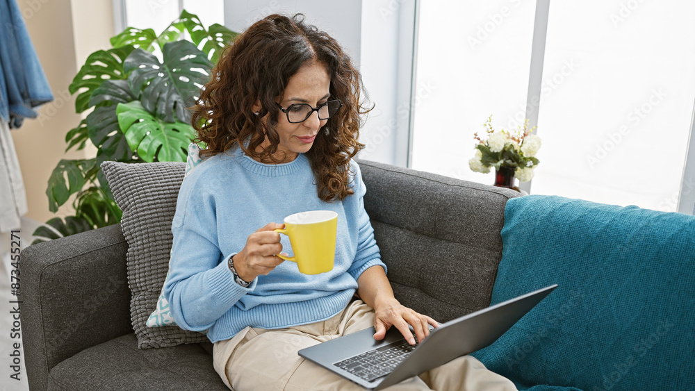 Sticker Hispanic woman enjoys coffee while working on laptop in cozy living room interior.