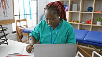 A focused african woman with braids taking notes in a physiotherapy clinic room.