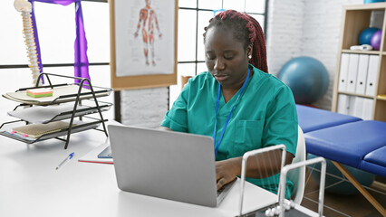 African american woman in scrubs using laptop at physiotherapy clinic center indoor workplace.