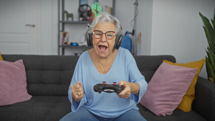Elderly woman excitedly playing video games with headphones on a sofa indoors.