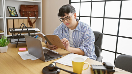 A young man reading a book in a modern office with a laptop, headphones, and plants in the background.