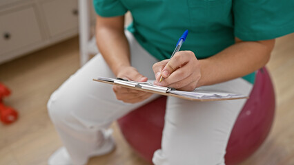Man in green scrubs sits on a balance ball indoors writing notes in a clinic setting