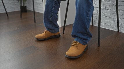 A man wearing blue jeans and casual brown shoes sits in an indoor waiting room with a wooden floor.