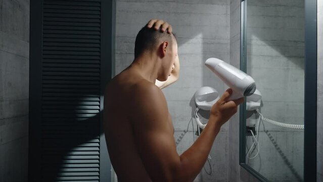 A worried young white man looks at himself in the mirror and inspects his premature receding hairline. Attractive Caucasian male adult in his 20s concerned about losing hair. Male pattern baldness. 