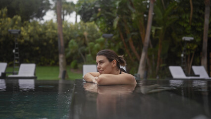 Fototapeta premium Young hispanic woman enjoying a serene moment in an outdoor pool at a bali resort, surrounded by lush tropical greenery.
