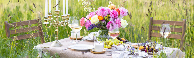 Elegant wedding or private party for a loving couple table setting outdoors in the garden. Beautiful decor with fresh ranunculi flowers, candles. Gourmet cheese board, fruits, white wine. Banner