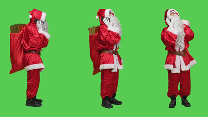 Modern santa claus answers phone call in studio with full body greenscreen, talking on remote chat using smartphone. Father christmas carries sack of presents and toys for holidays.