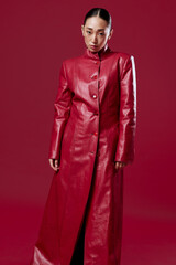Fashionable asian woman in a red leather coat posing with hands on hips against a red background, with hair in a ponytail
