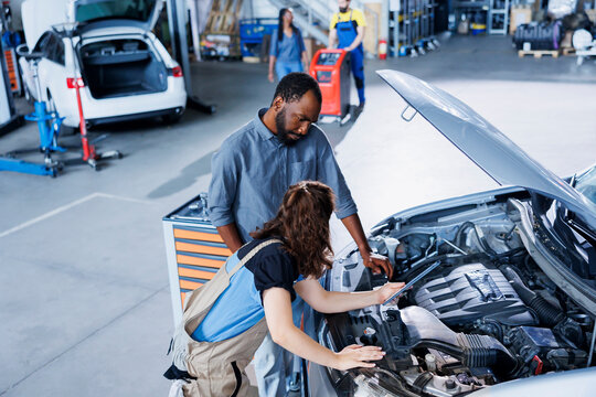 Skillful mechanic helping client with car checkup in auto repair shop. Employee in garage facility looking over automobile parts with woman, mending her vehicle steering mechanism during inspection