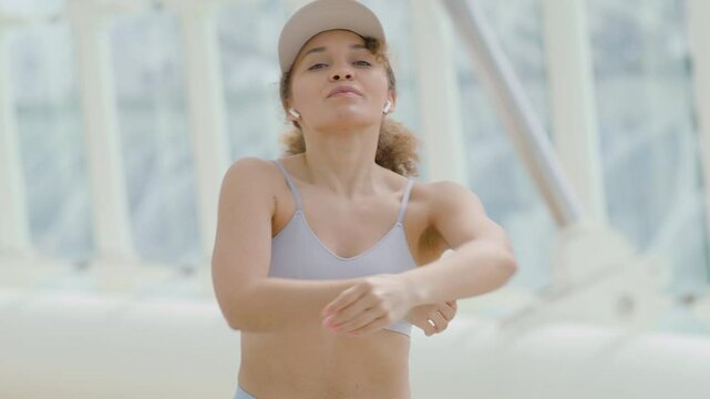 A focused young woman in a grey sports bra and cap is dedicated to her workout routine, doing stretching exercises indoors while wearing wireless earbuds, showcasing determination and concentration
