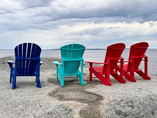Four blue, turquoise and red adirondack chairs perched on a huge rock overlooking the ocean on a...