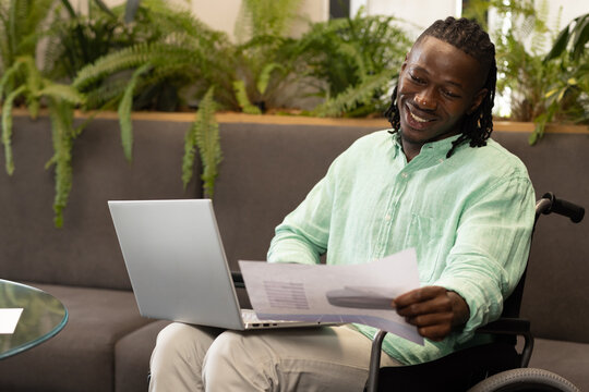Smiling man in wheelchair working on laptop and reviewing documents in office