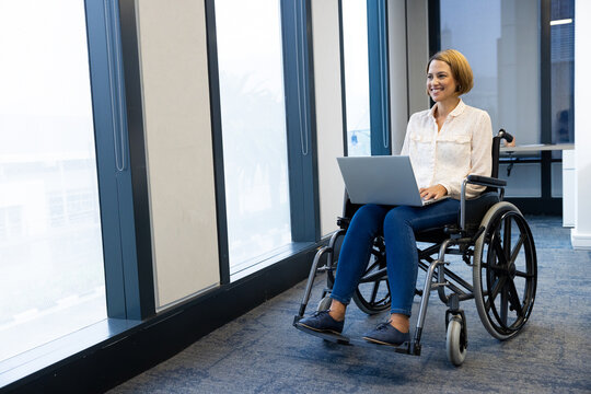 Smiling woman in wheelchair using laptop, working in modern office environment, copy space