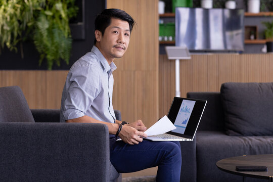 Working on laptop, man analyzing financial charts and holding documents in office