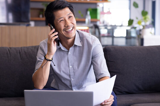 Smiling man holding paper and talking on smartphone, working on laptop