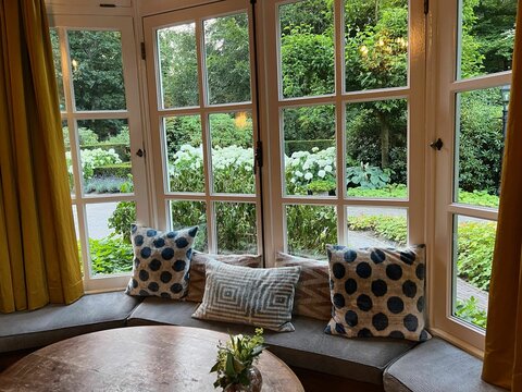 Wooden table near bay window seat with cushions in cozy room. Interior design