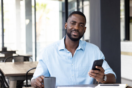 Holding smartphone and coffee cup, man sitting at table and smiling, copy space