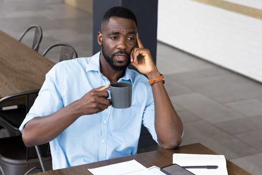 Holding coffee mug, man thinking while sitting at table with smartphone and papers