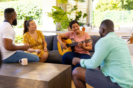 Playing guitar, woman entertaining diverse friends while sitting on couch at home gathering