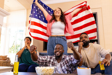 Celebrating with diverse friends, woman holding American flag while others cheer on couch