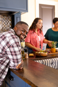 Smiling man leaning on kitchen counter while diverse friends preparing food together