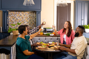 Diverse friends toasting drinks and enjoying meal together at home dining table