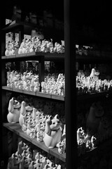 A black and white photo of shelves overflowing with Maneki-neko cat figurines of various sizes in Gotokuji Temple