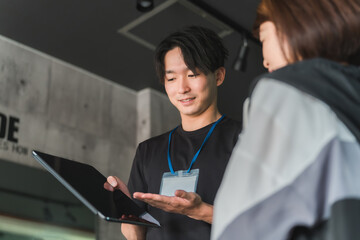 ジムで女性会員に運動メニューの説明・指導をするトレーナー・インストラクターの若いアジア人男性
