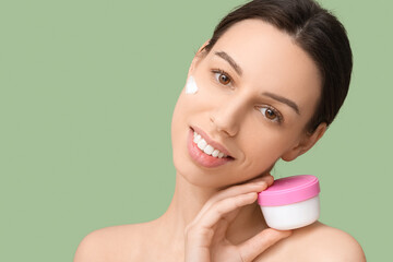 Beautiful young woman with cream on cheek holding jar of sunscreen against green background